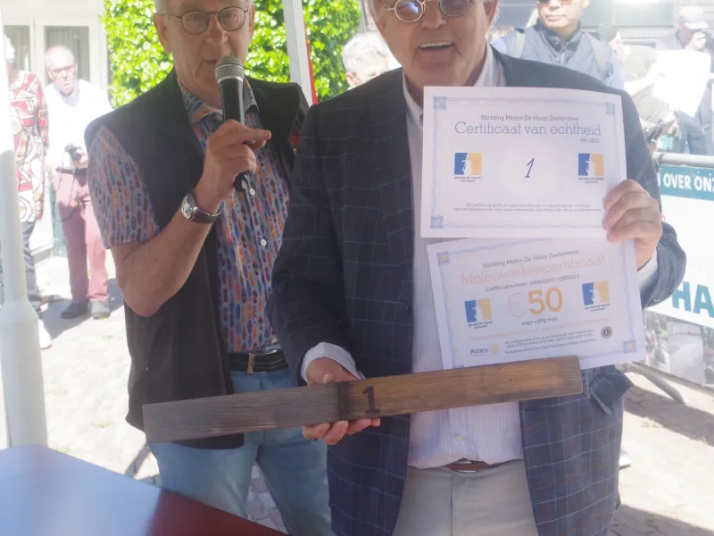 Twee heren staan lachend op de foto. Een ervan houdt een certificaat en een stuk heklat van de molen vast.